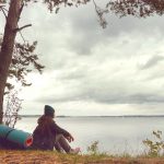 Traveling lonely woman sitting near forest lake and looking far away