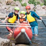 Young family of three wearing life vests and helmets posing beside rowing catamaran