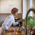 A young woman with cup of coffee standing indoors in kitchen, looking at plant.