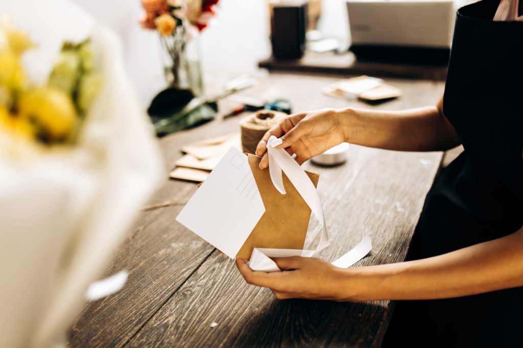 Florist is packing a postcard in an envelope with white ribbon on the wooden table