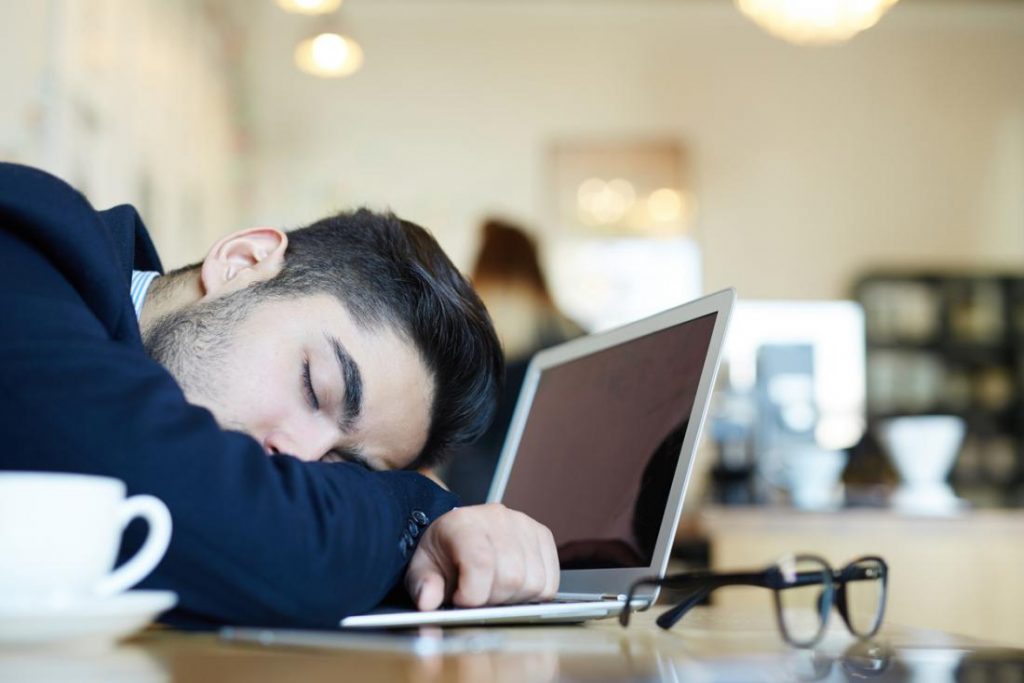 man asleep at desk in front of laptop and coffee cup
