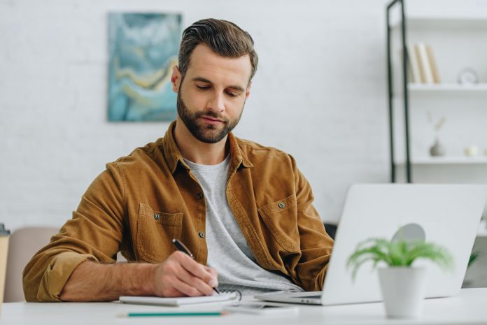 handsome man writing in notebook with pen in apartment