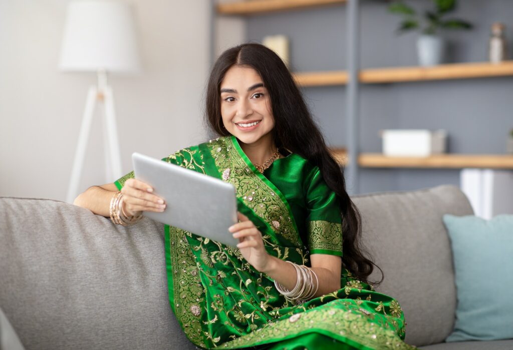 Cheerful Indian lady in traditional saree sitting on cozy sofa with tablet pc attending online