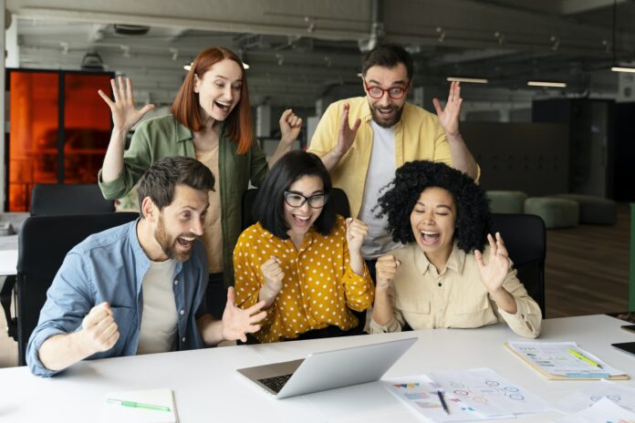 Team of diverse business people clench fists winning tender, sitting at desk with laptop and graphs